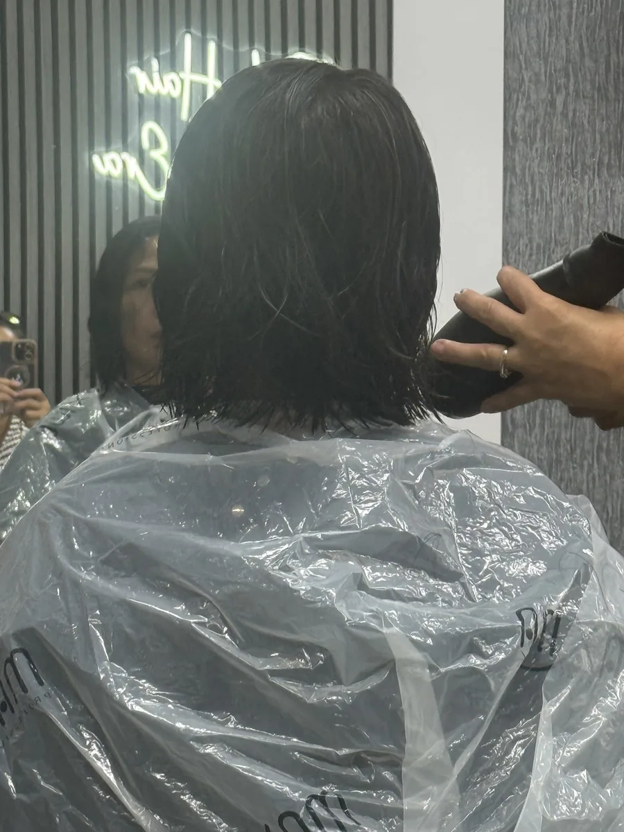 Stylist's hands working through a dark bob mid-treatment, client gowned in a clear cape