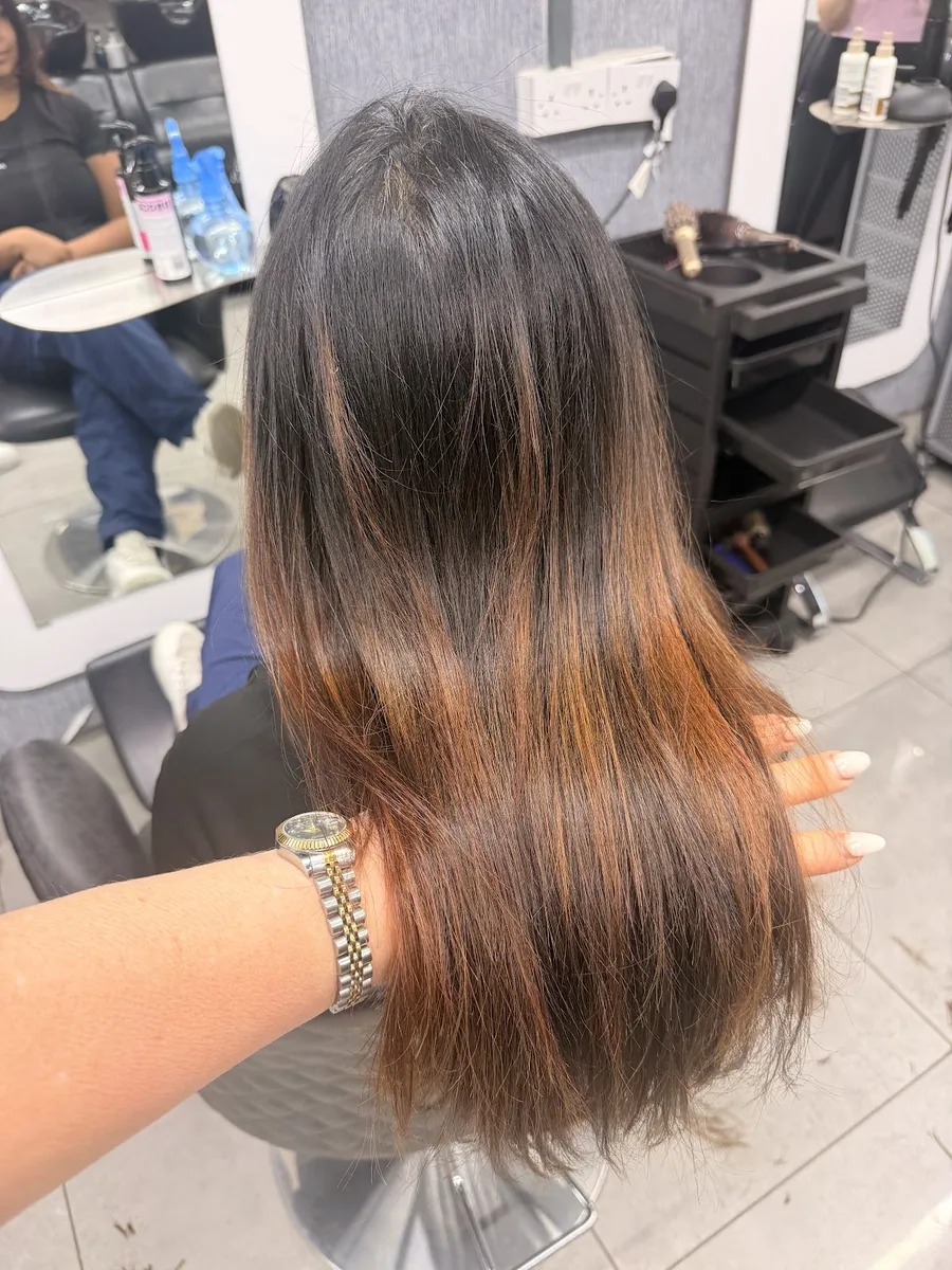 Stylist's hand lifting a section of long dark hair with a copper ombre running through the lengths